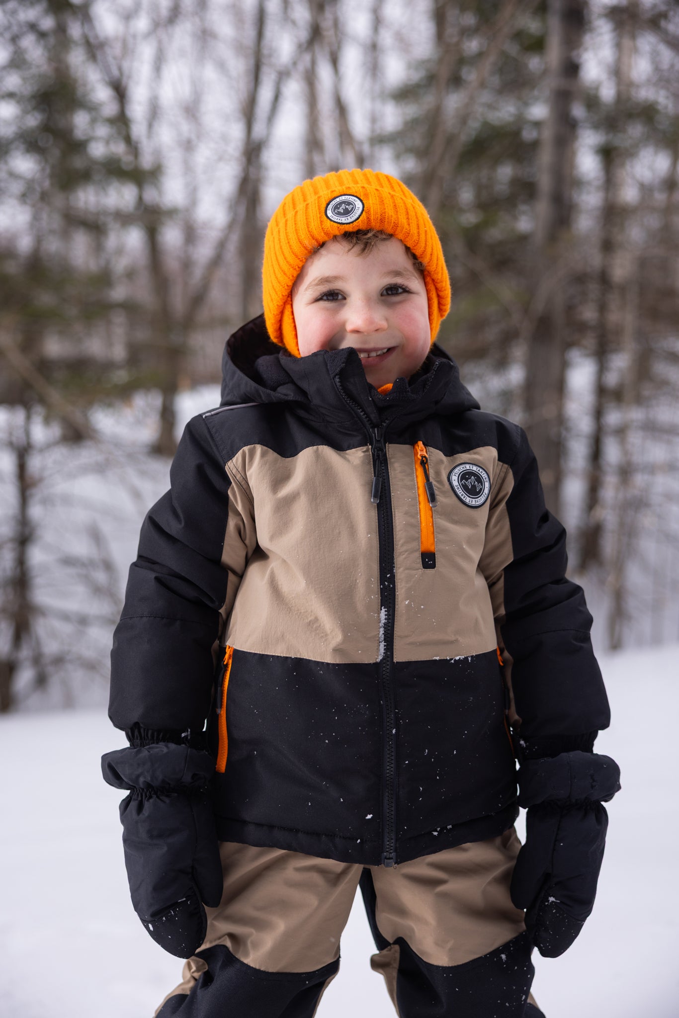 Habit de neige - Peluche & Tartine enfant beige et noir avec détails orange, en tissu isolant, porté par un garçon souriant en extérieur, vue de face.