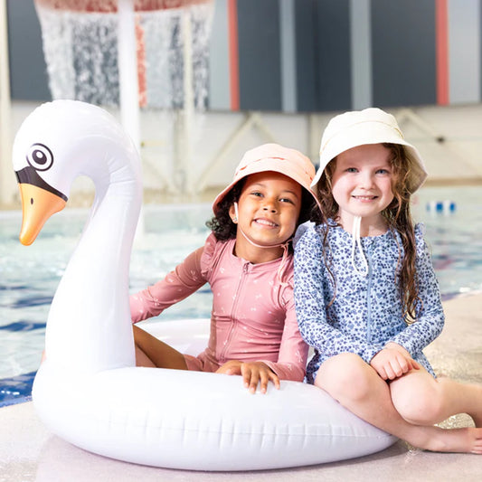 Deux filles portant des Chapeau U.V - Nanö beige et rose, assises sur un flamant rose gonflable, bord de piscine, vue de face.