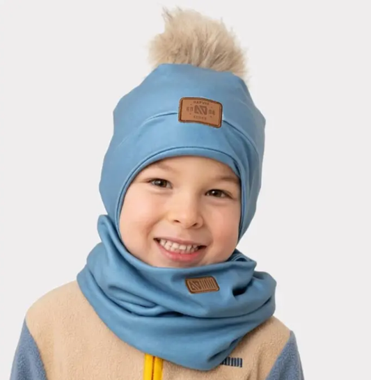 Enfant souriant portant une Tuque d'hiver - Nanö bleue avec pompon beige en fausse fourrure, ensemble cache-cou assorti, vue de face.