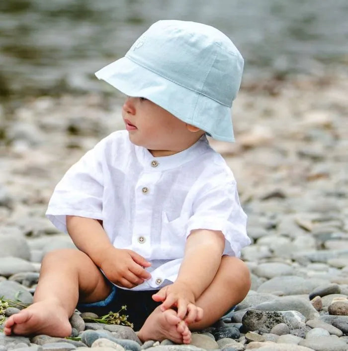 Chapeau en lin - Calikids bleu clair en lin, sangles velcro, pour bébé garçon, porté assis de profil sur décor naturel.