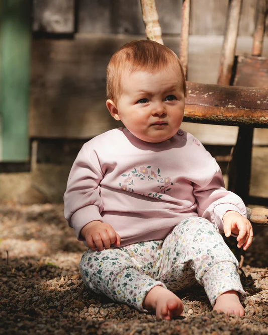 Ensemble - Deux Par Deux pour bébé fille, sweat rose à motif floral et pantalon blanc imprimé fleurs, assise en extérieur, vue de face.