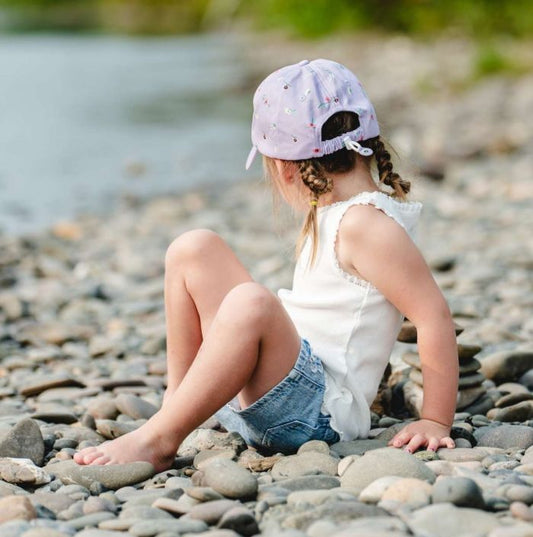 Casquette - Calikids violette à motifs, coton, ajustable à l'arrière, portée par enfant, vue de dos assise sur des cailloux.