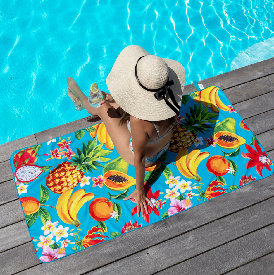 Serviette de plage - L’île aux saveurs à motifs fruités multicolores sur fond bleu, microfibre, vue de dessus avec femme au bord de piscine.
