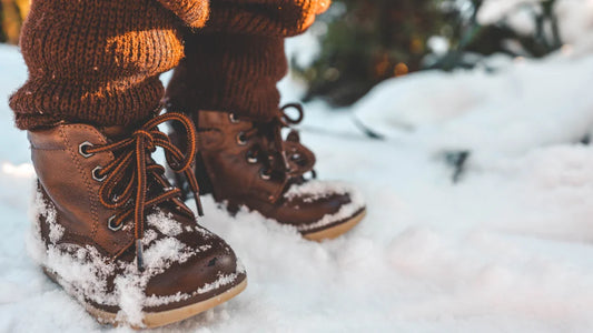 Un enfant portant des bottes d'hiver marron joue dans la neige, entouré d'un paysage enneigé.