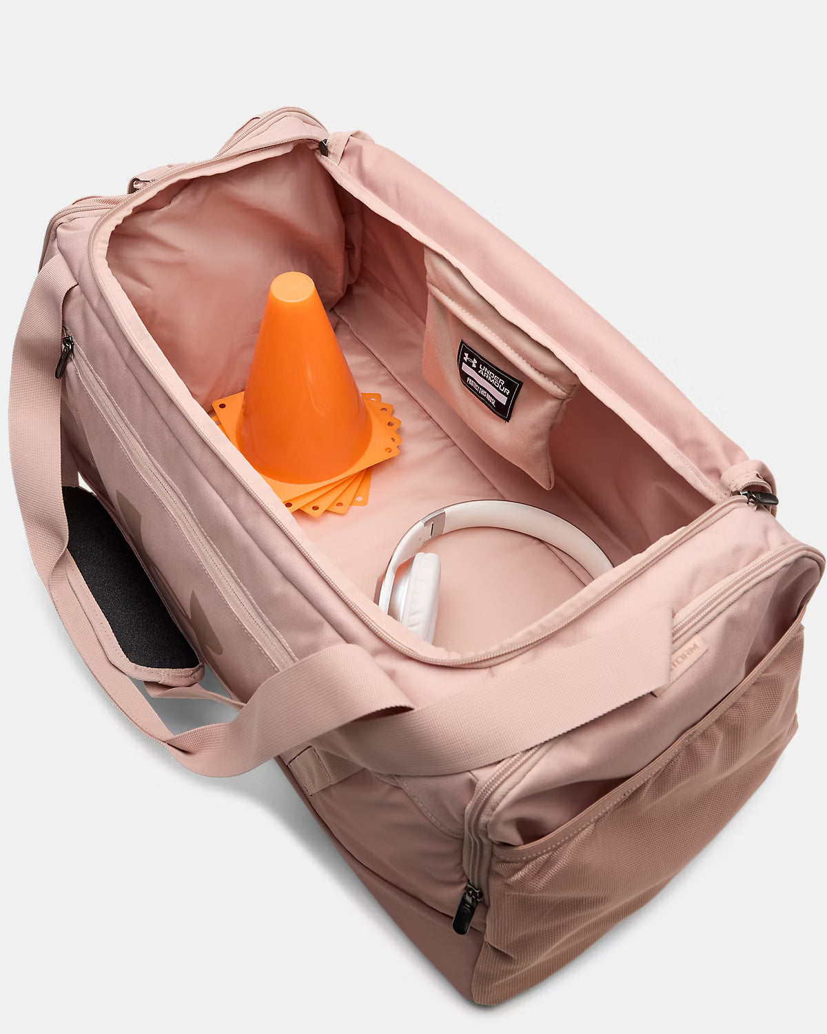 Sac sport - Under Armour rose clair en tissu synthétique, vue de dessus avec ouverture principale, contenant un casque blanc et un cône orange.
