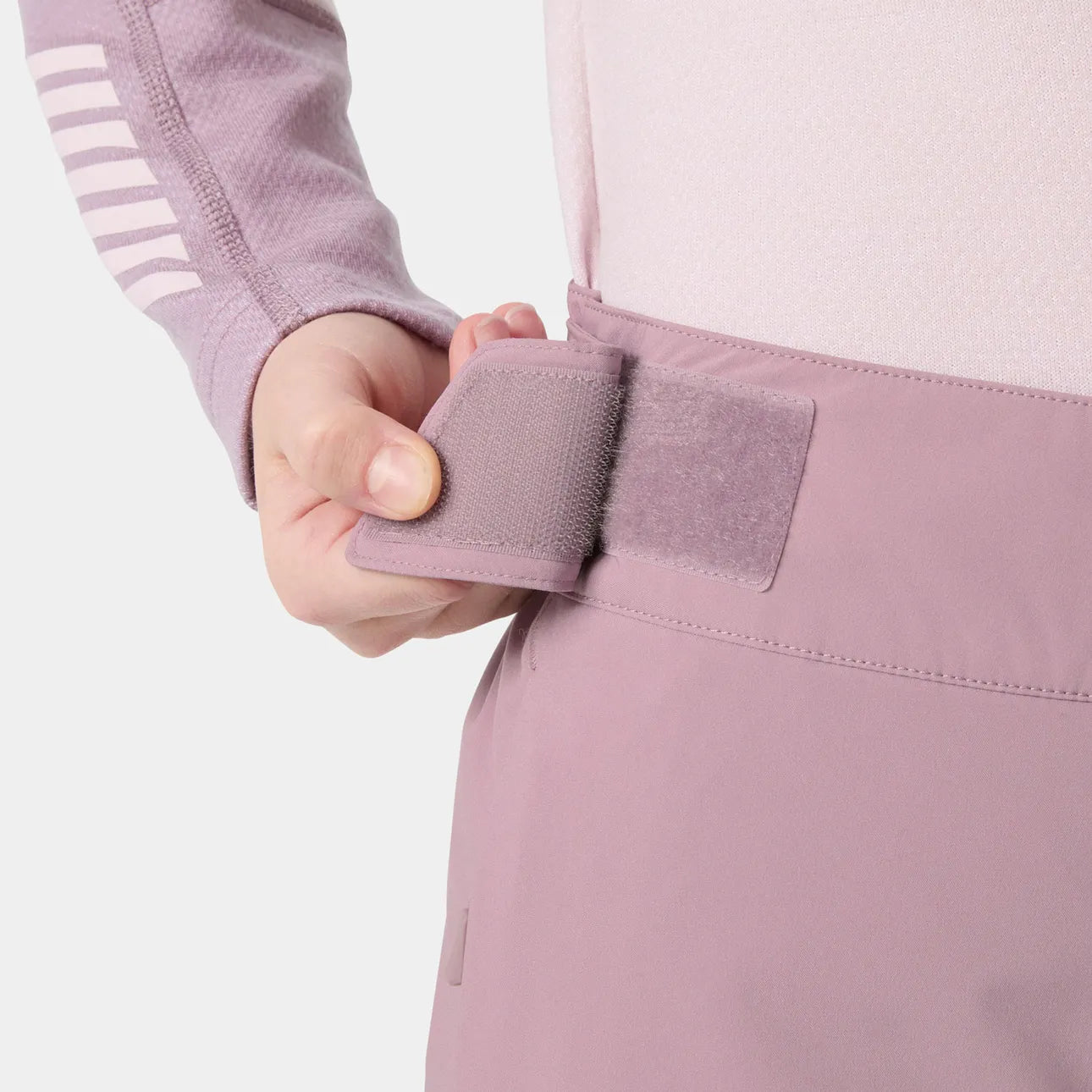 Détail d'une main ajustant une bande auto-agrippante mauve sur la taille d'un pantalon technique rose clair, vue rapprochée côté gauche.