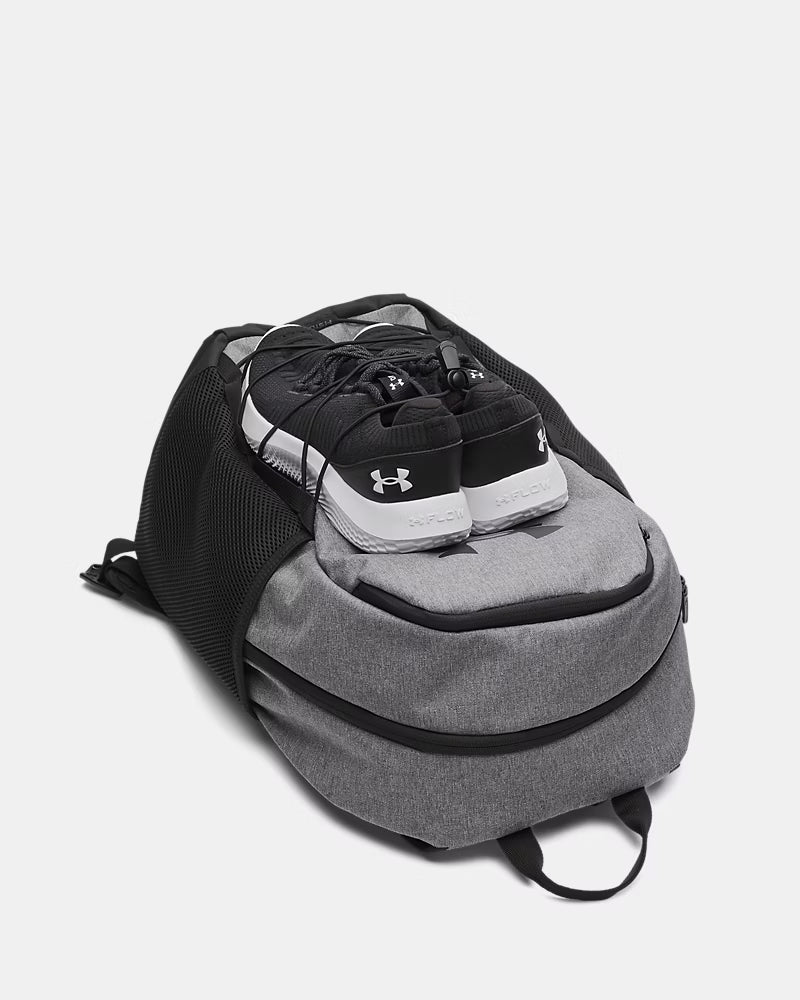 Sac à dos UA Hustle Sport 6.0 - Under Armour (Copie) gris et noir en polyester, style sport, avec chaussures noires posées sur le dessus, vue de face légèrement plongée.