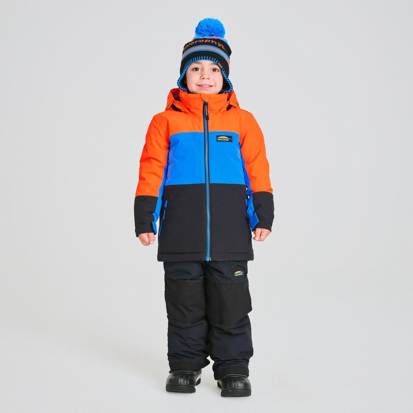 Habit de neige Colibri - Chlorophylle avec manteau tricolore orange, bleu et noir, pantalon noir, porté par enfant, vue de face.