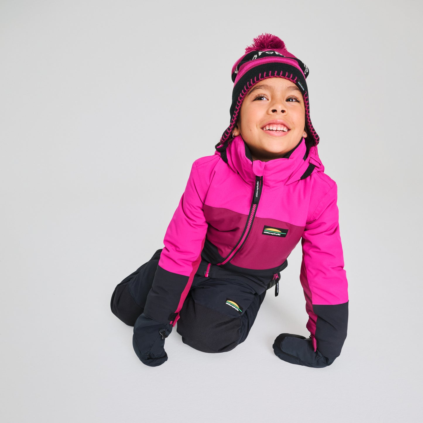 Habit de neige Colibri - Chlorophylle rose et violet foncé en tissu technique, pour enfant, porté de face en position agenouillée avec bonnet assorti.