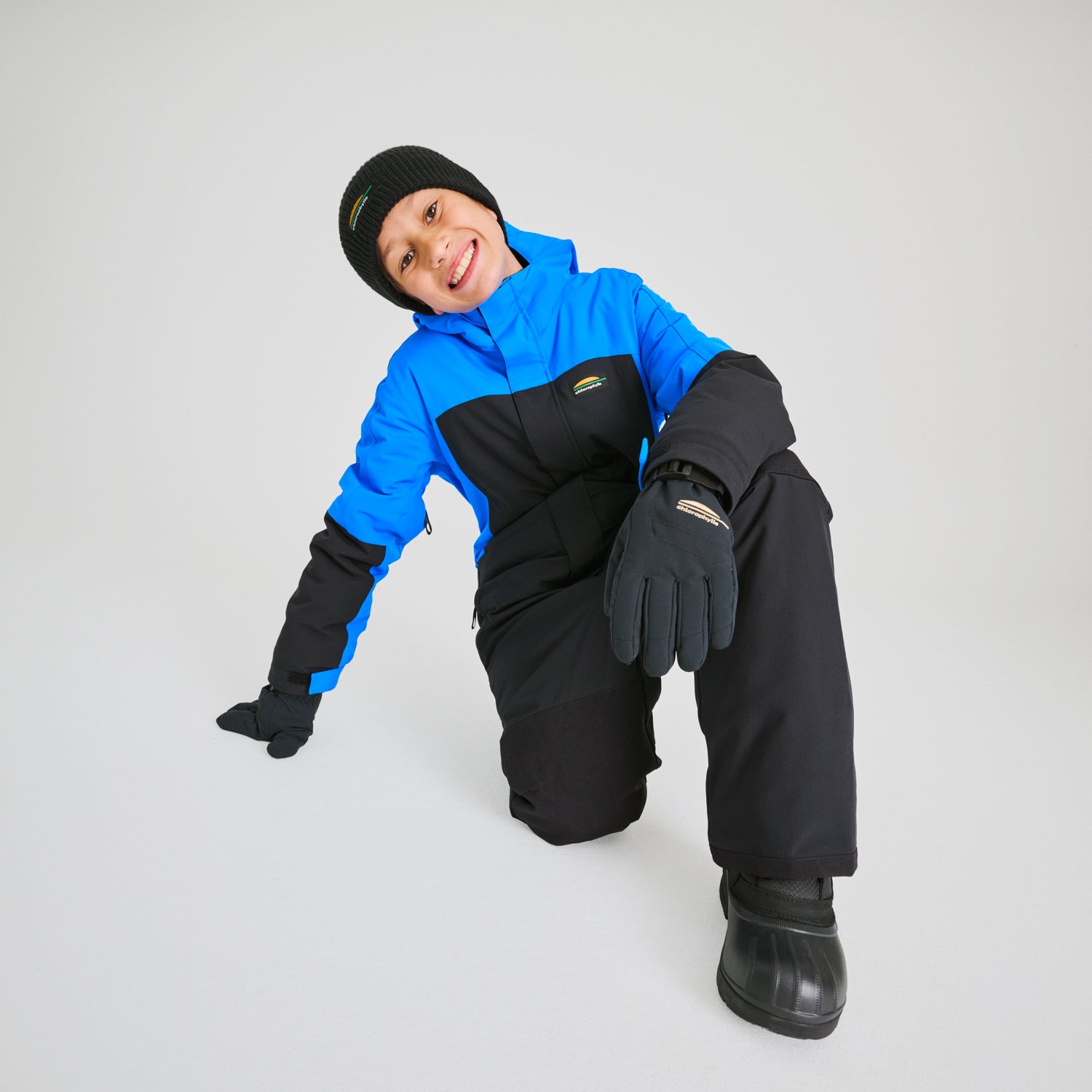 Habit de neige 1 Pièce - Chlorophylle bleu et noir en textile isolant, pour enfant, porté assis de face avec bonnet et gants noirs.