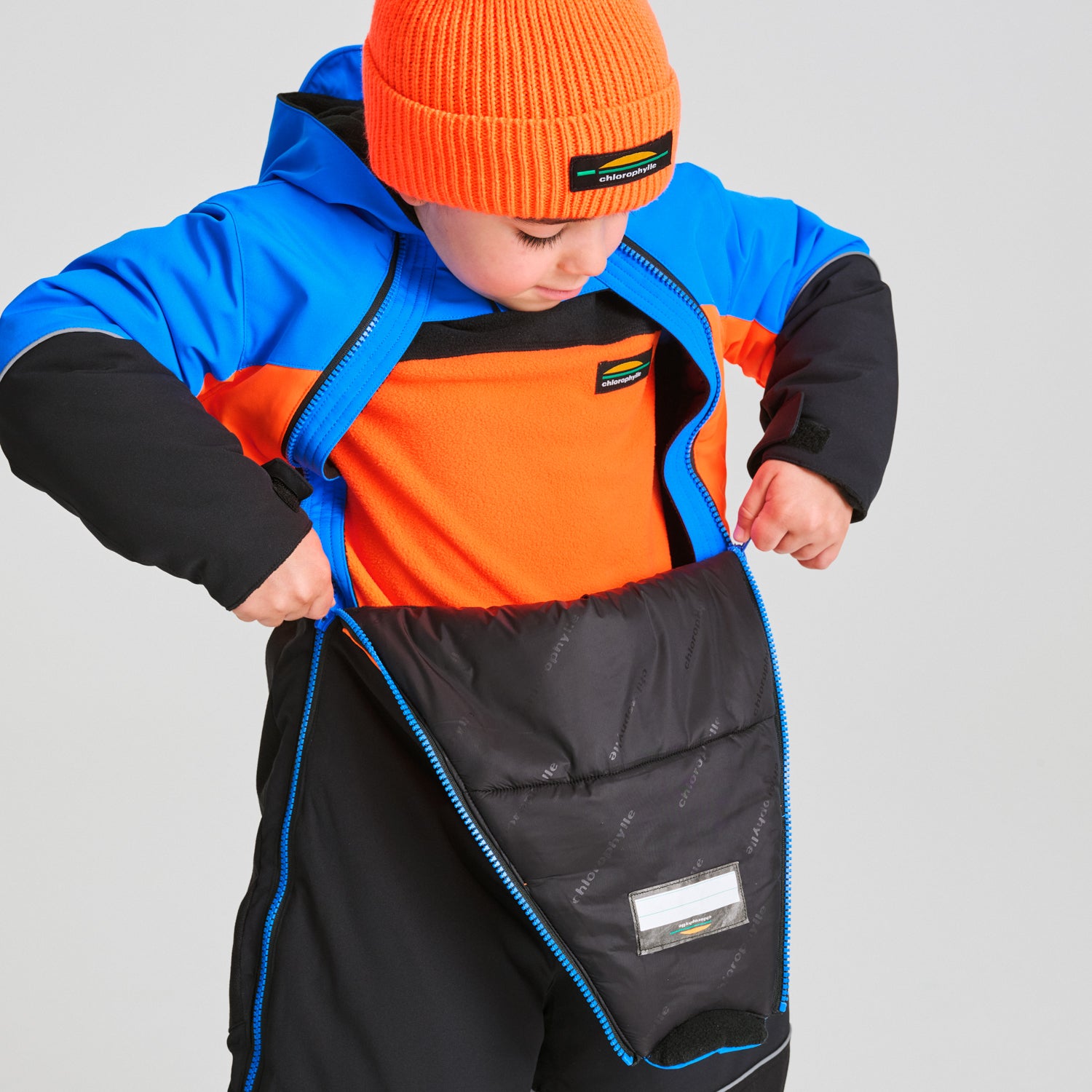 Enfant portant un Habit de neige 1 pièce - Chlorophylle bleu, noir et orange, en train de zipper la combinaison, vue de face avec capuche et bonnet orange.