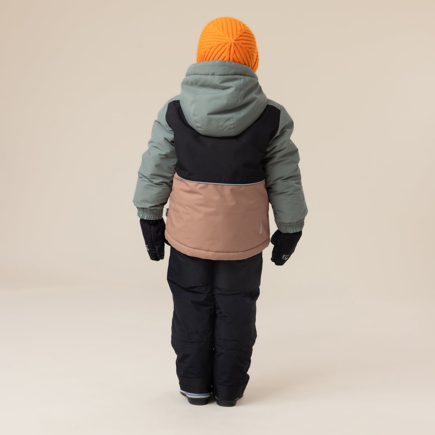 Enfant portant habit de neige - Peluche & Tartine, combinaison trois tons beige, noir et vert kaki, bonnet orange, gants noirs, vue de dos.