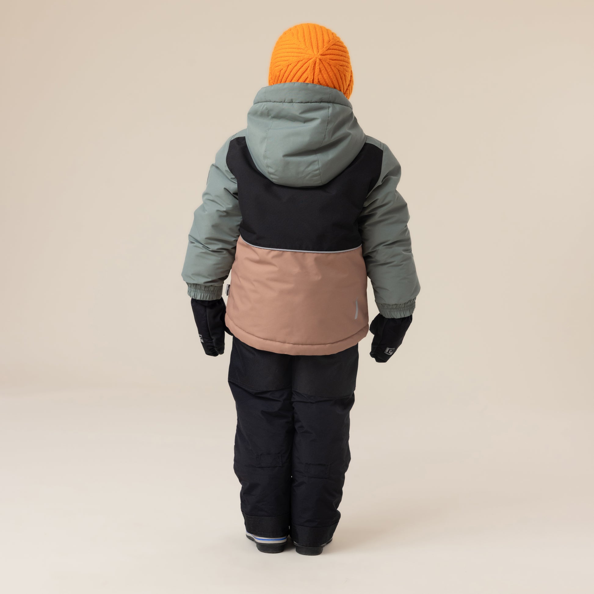 Enfant portant habit de neige - Peluche & Tartine, combinaison trois tons beige, noir et vert kaki, bonnet orange, gants noirs, vue de dos.