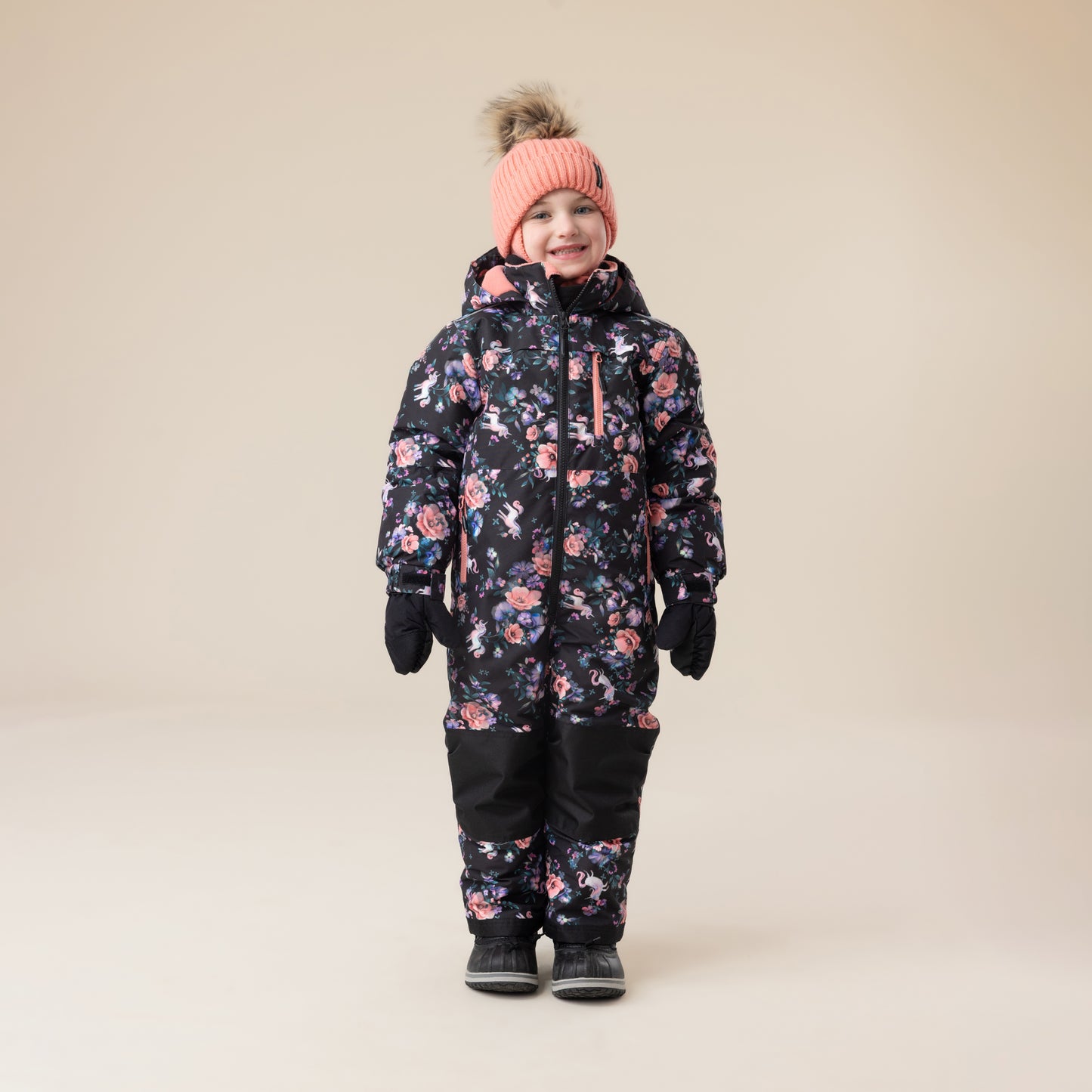 Habit de neige 1 Pièce - Peluche & Tartine, combinaison noire à motifs floraux roses et mauves, pour enfant, vue de face portée par un garçon avec bonnet rose et moufles noires.
