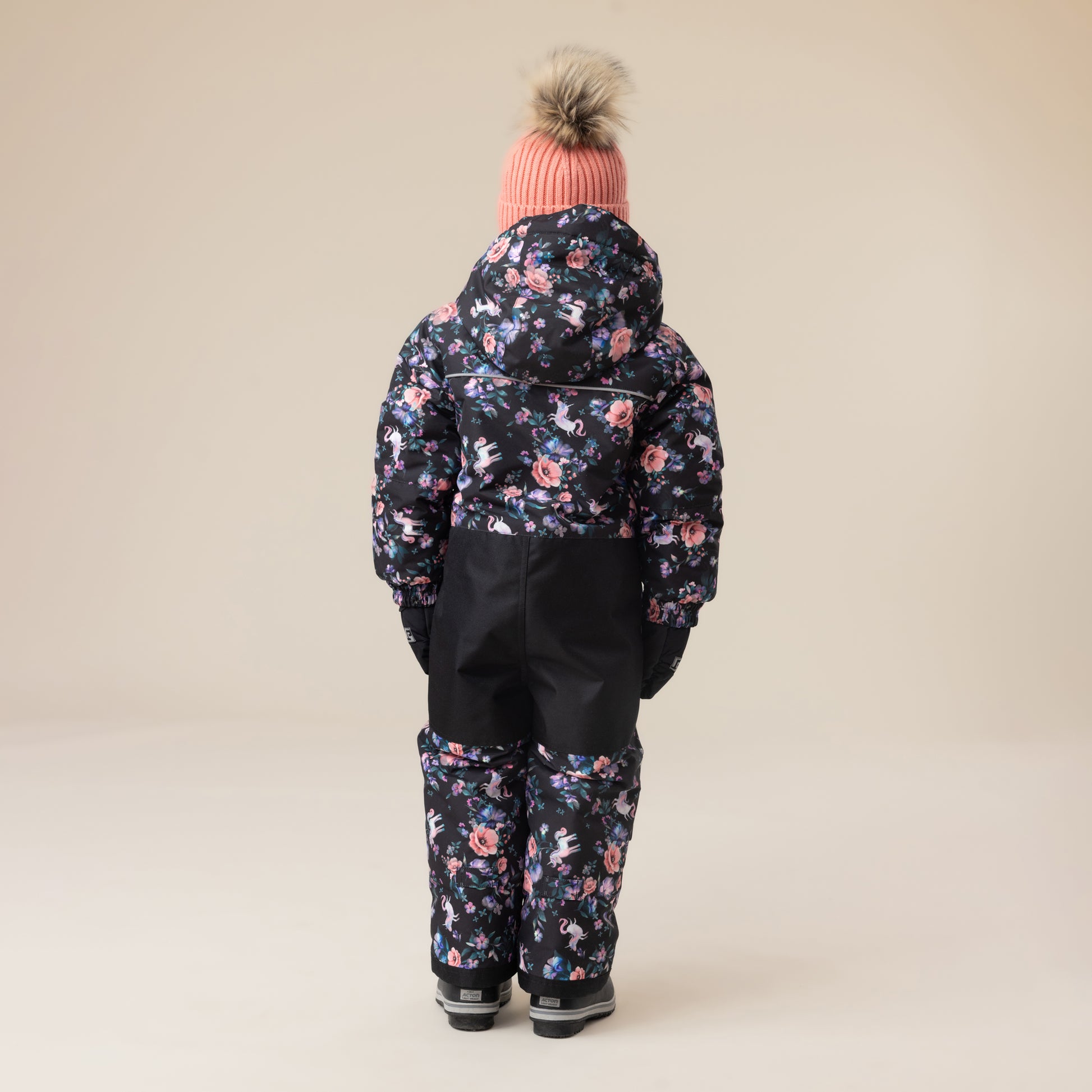 Habit de neige 1 Pièce - Peluche & Tartine pour enfant, combinaison noire avec imprimé floral rose et bleu, vue de dos portée avec bonnet rose à pompon.