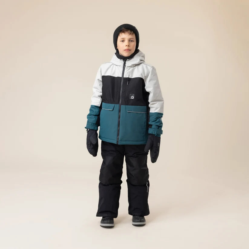 Garçon portant une tenue de ski noire bleu pétrole et blanche avec moufles assorties vue de face dans un studio beige