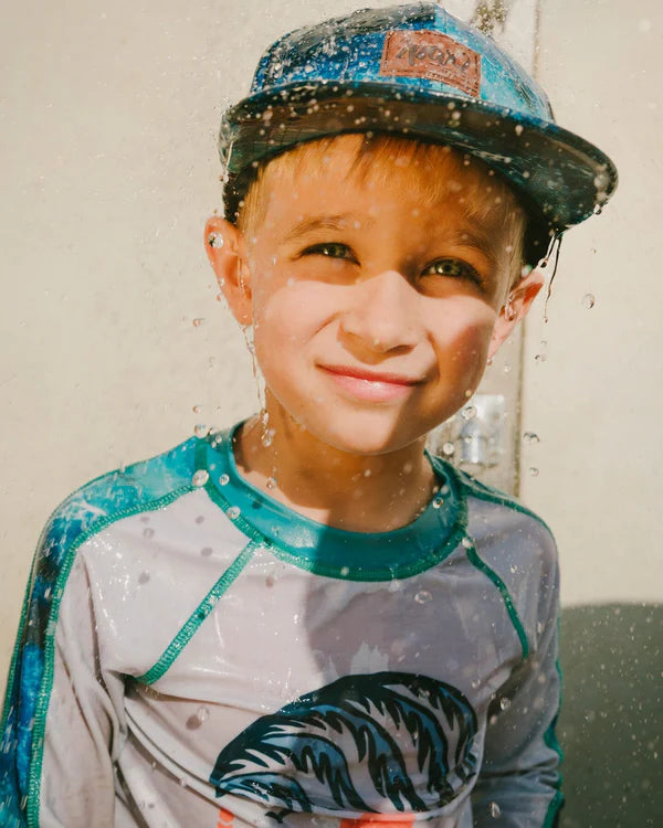 Casquette - Deux Par Deux bleu à imprimé vagues, visière plate, portée par garçon sous jets d'eau, vue de face.