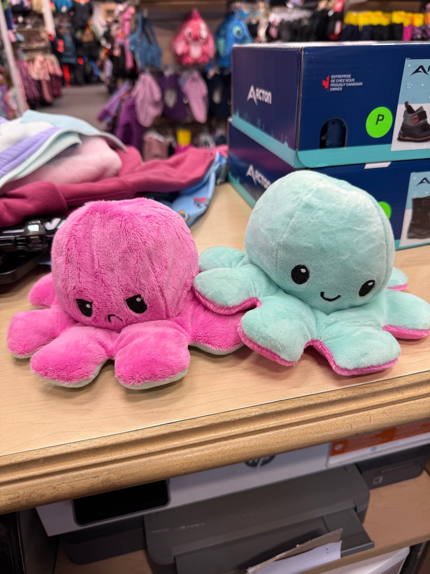Pieuvre En Peluche Réversible en tissu doux rose et turquoise, expressions joyeuse et contrariée, vue de face sur table en magasin.