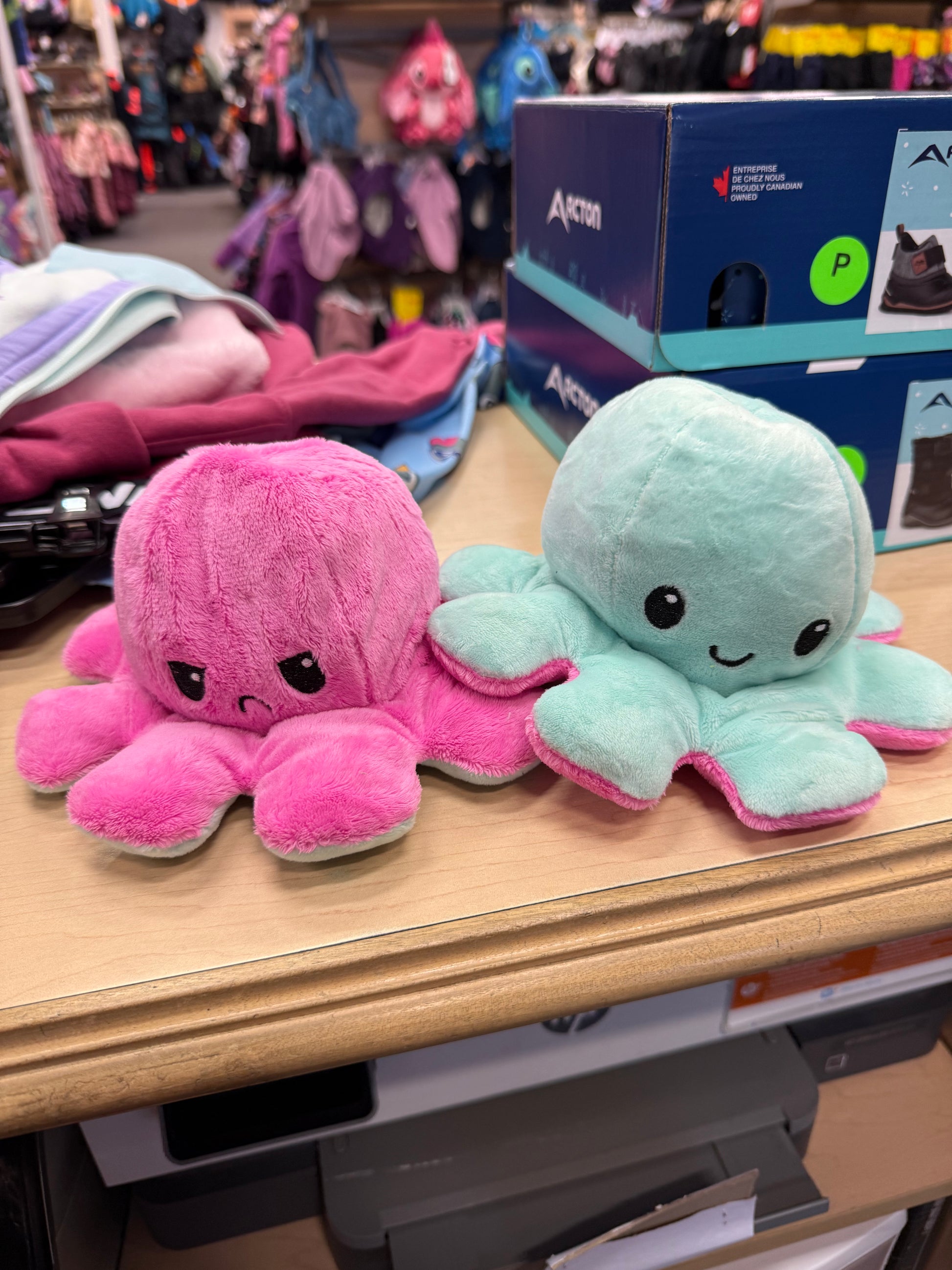 Pieuvre En Peluche Réversible en tissu doux rose et turquoise, expressions joyeuse et contrariée, vue de face sur table en magasin.