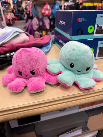Pieuvre En Peluche Réversible en tissu doux rose et turquoise, expressions joyeuse et contrariée, vue de face sur table en magasin.