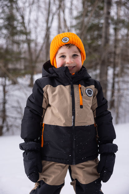 Habit de neige - Peluche & Tartine enfant beige et noir avec détails orange, en tissu isolant, porté par un garçon souriant en extérieur, vue de face.