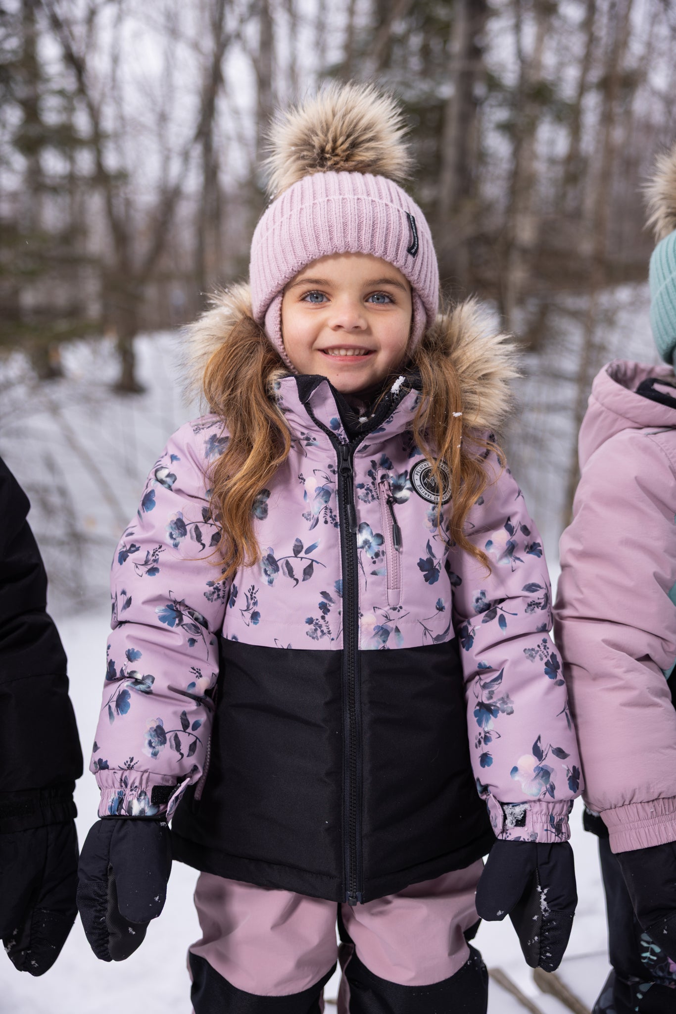 Enfant fille portant un Habit de neige - Peluche & Tartine violet à motifs floraux bleus et noirs avec capuche à fourrure, bonnet rose à pompon, vue de face en extérieur hiver.