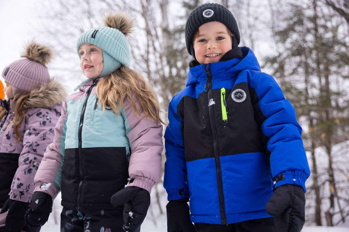 Enfants portant des habits de neige - Peluche & Tartine, manteau bleu et noir avec fermeture éclair verte sur garçon et manteau rose, mauve et vert clair sur fille, en extérieur neigeux, vue de face.