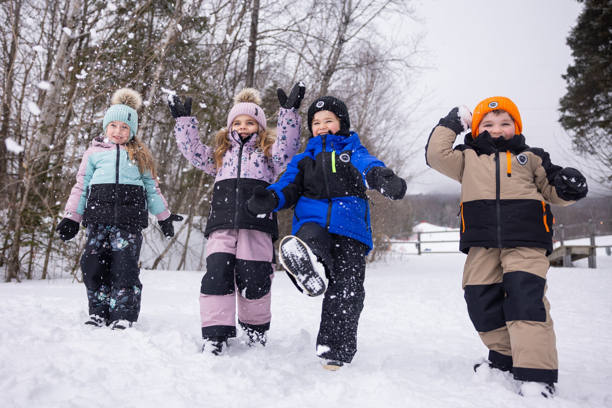 Quatre enfants portant des habits de neige - Peluche & Tartine dans les tons bleu, rose, beige et noir, jouant dans la neige, vue en pied de face.