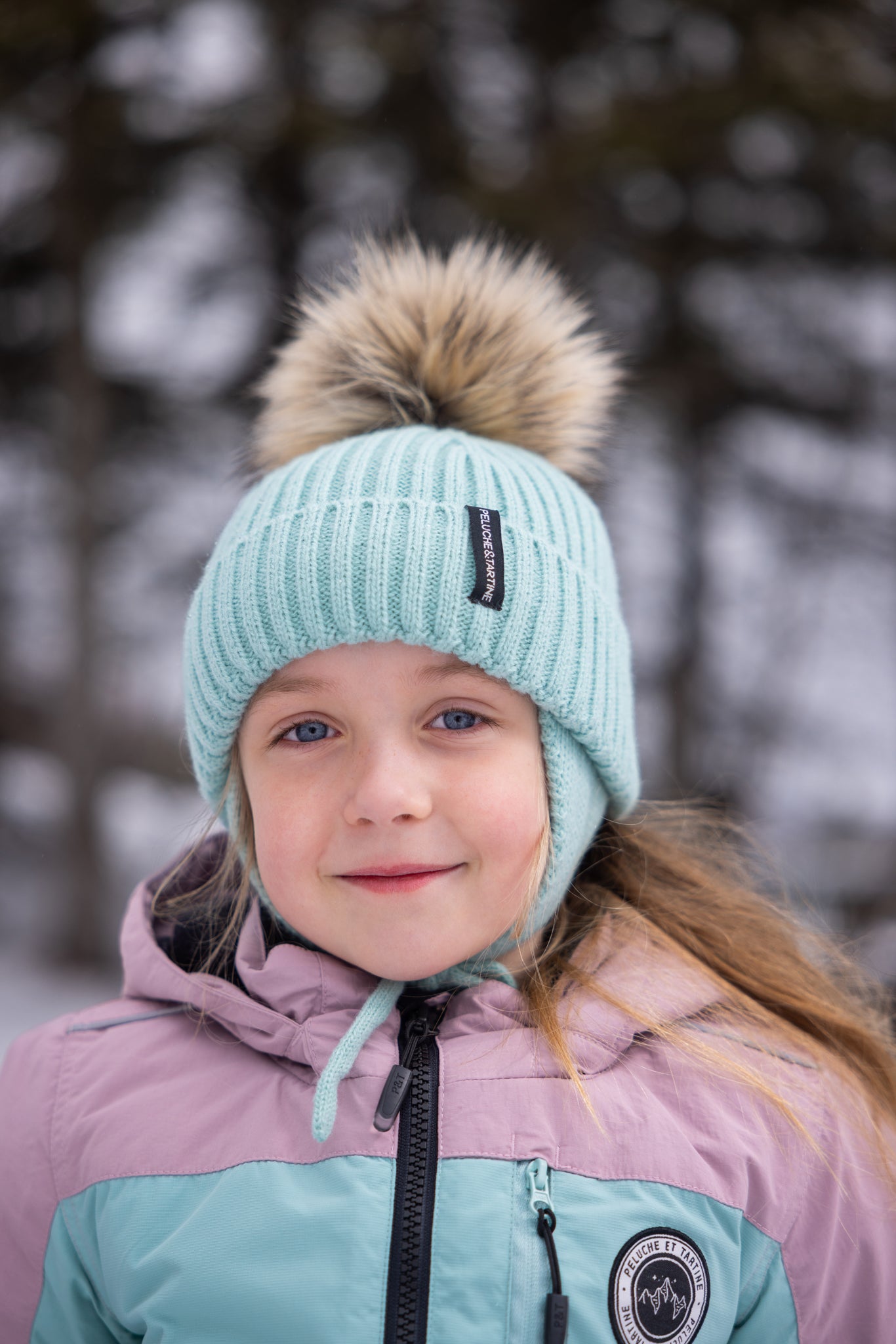Fillette portant un habit de neige - Peluche & Tartine en tissu imperméable mauve et turquoise, vue de face, bonnet tricoté bleu ciel avec pompon en fausse fourrure.