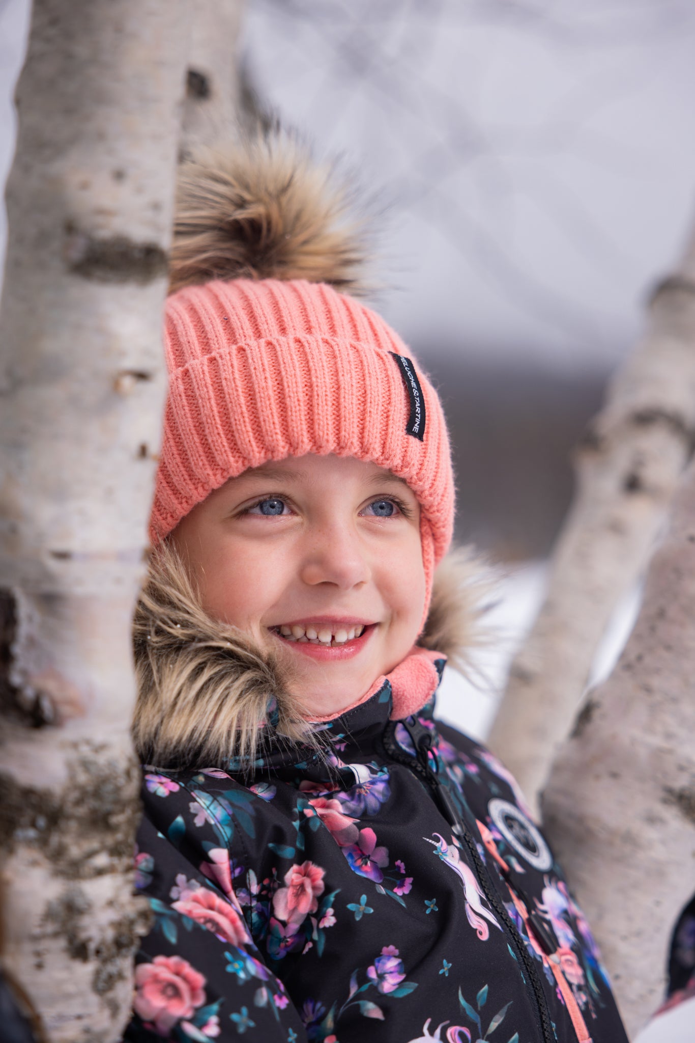 Enfant souriant portant un habit de neige - Peluche & Tartine noir à motifs floraux roses et bleus, bonnet rose avec pompon en fausse fourrure, vue de face.