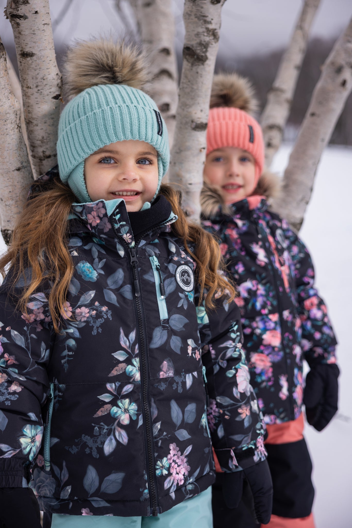 Habit de neige - Peluche & Tartine noir à motifs floraux rose, bleu et violet, porté par deux filles enfants, bonnet à pompon en maille, vue de face.