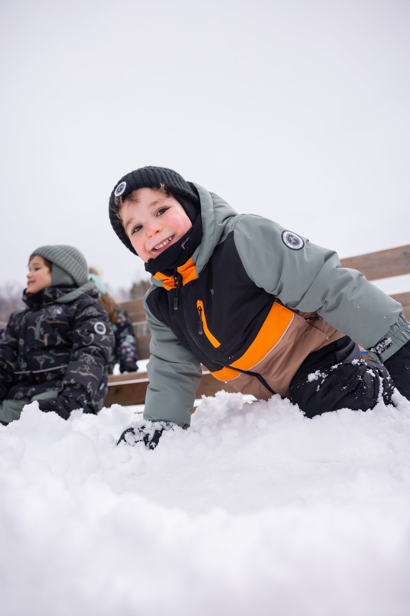 Habit de neige - Peluche & Tartine gris, noir et orange, en matière imperméable, porté par un enfant garçon jouant dans la neige, vue de face.