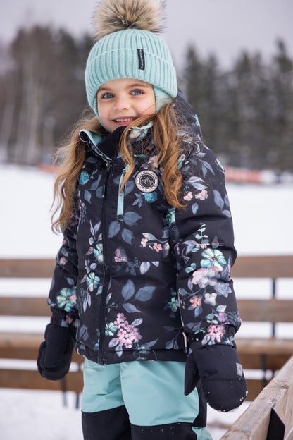 Habit de neige - Peluche & Tartine pour enfant fille, veste noire à motifs floraux bleu, rose et vert, vue de trois quarts portée avec un bonnet bleu clair à pompon.