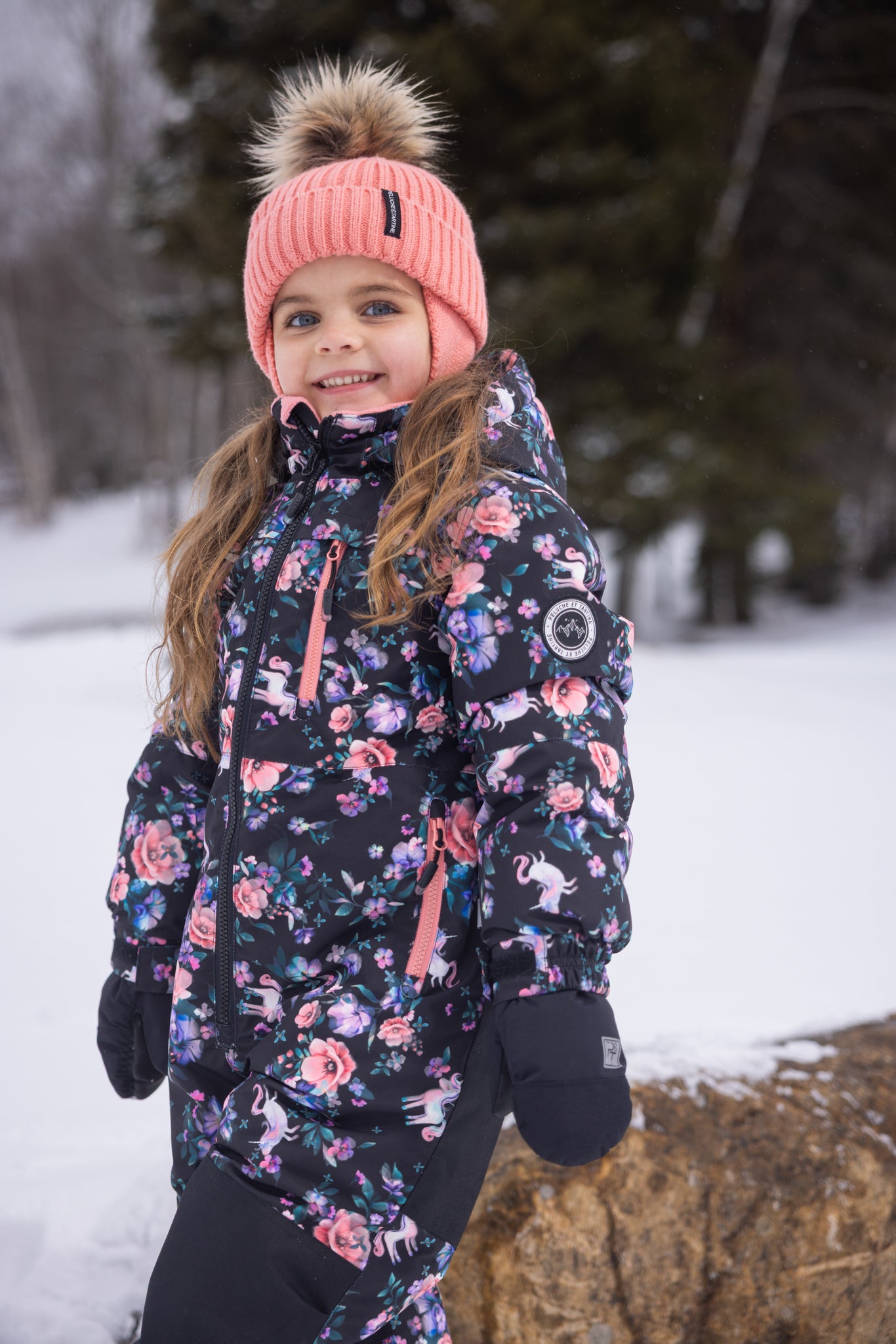 Habit de neige 1 Pièce - Peluche & Tartine à imprimé fleuri rose et violet sur fond noir, en tissu isolant, porté par une enfant souriante avec bonnet rose à pompon, vue de côté.