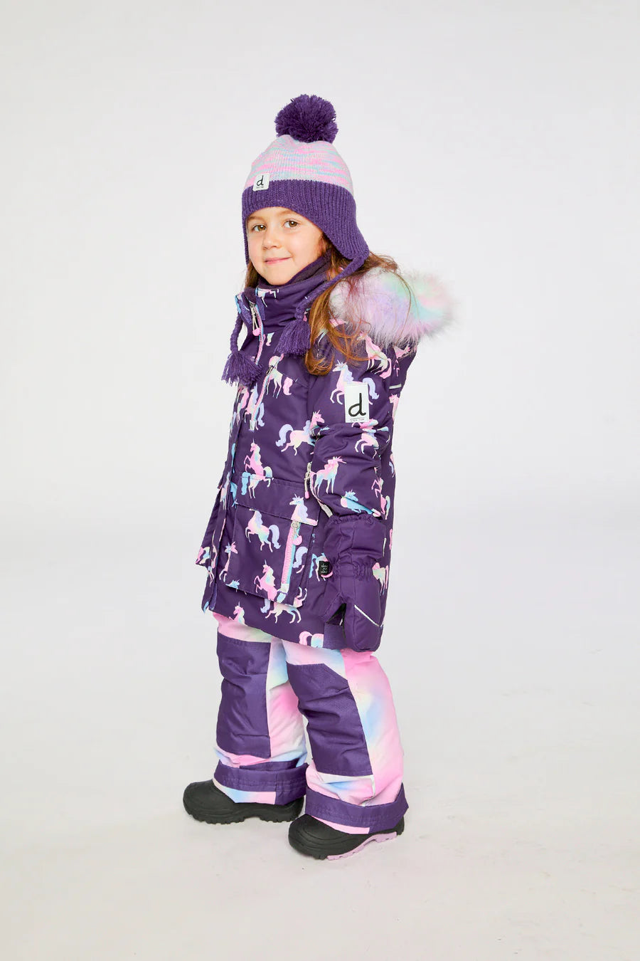 Habit de neige - Deux Par Deux violet à motifs licornes roses et bleues, avec capuche à fourrure colorée, pantalon pastel, porté par enfant fille, vue de profil.