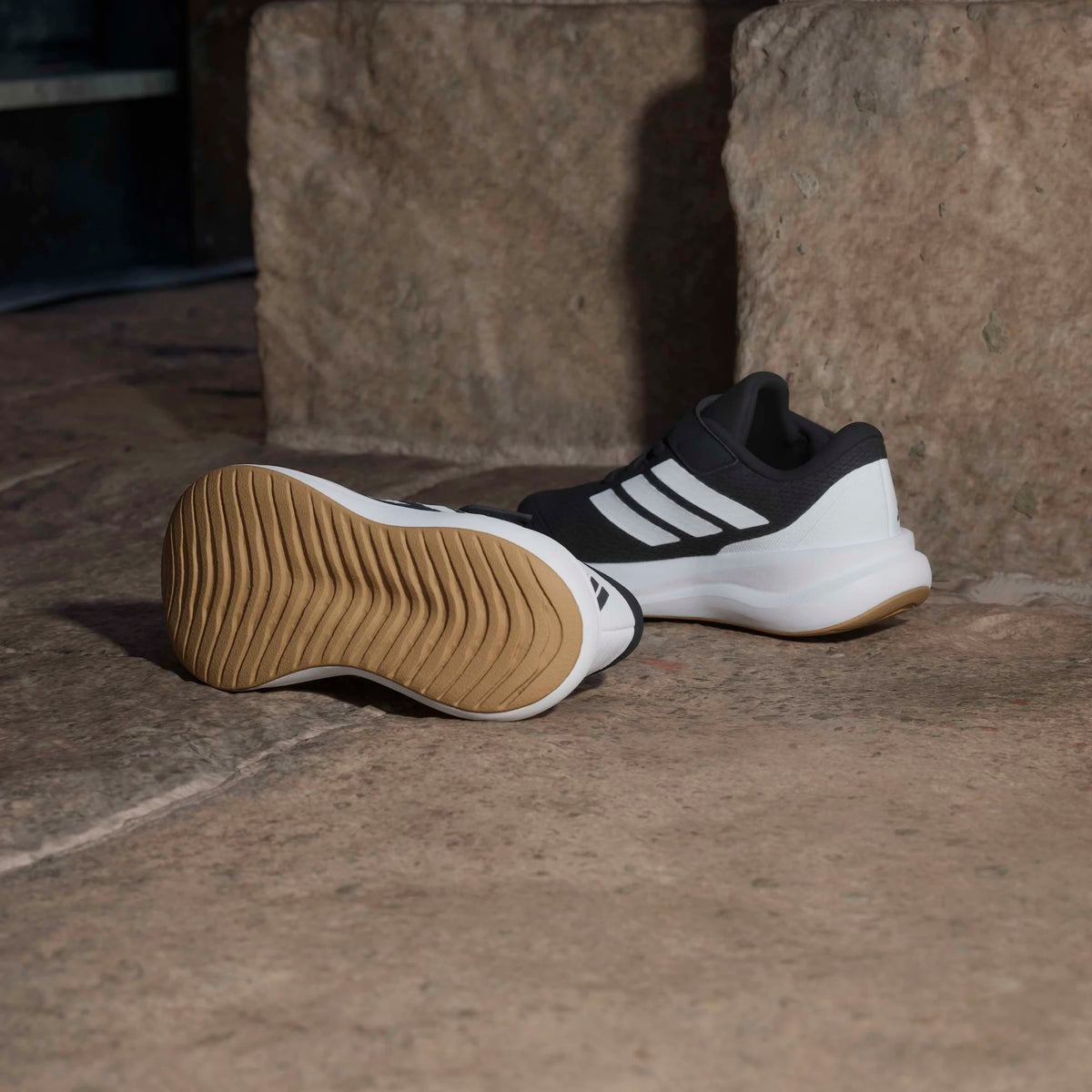 Paire de baskets de sport noires et blanches posées au sol avec semelle extérieure beige visible et vue latérale