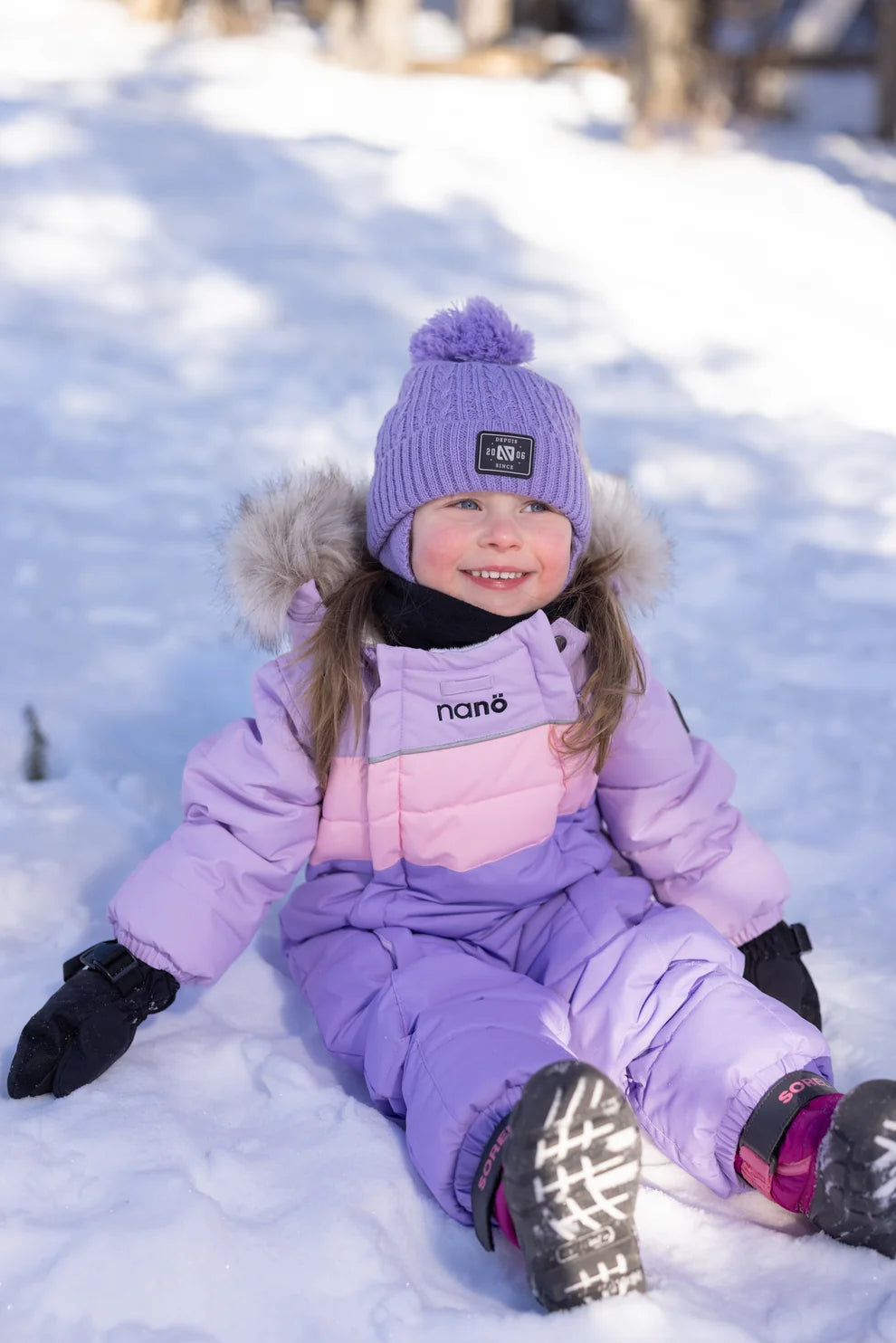 Habit de neige Lyvia - Nanö violet et rose pâle avec capuche bordée de fausse fourrure, porté par une enfant assise dans la neige, vue de face.