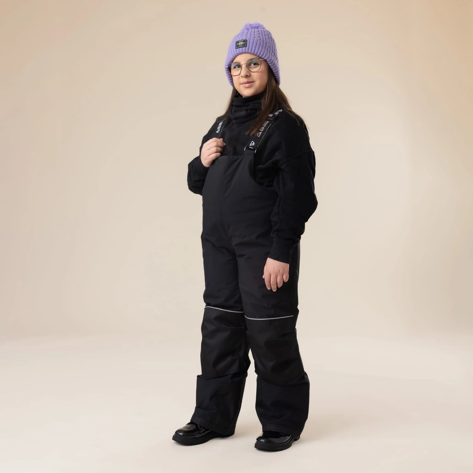 Salopette de neige Maxifit - Nanö noire en matériau imperméable, portée par une enfant fille, vue de face, avec un bonnet violet et un pull noir.