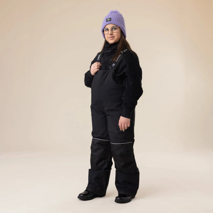 Salopette de neige Maxifit - Nanö noire en matériau imperméable, portée par une enfant fille, vue de face, avec un bonnet violet et un pull noir.