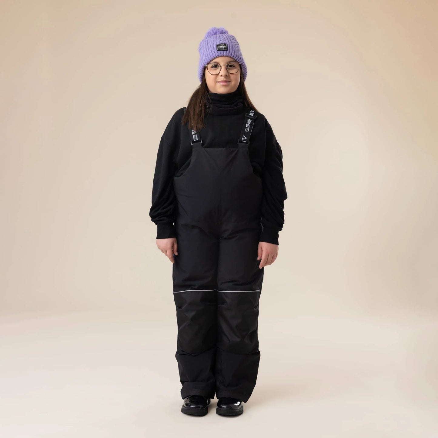 Salopette de neige Maxifit - Nanö noire, tissu imperméable avec renforts aux genoux, portée par enfant fille vue de face, style décontracté hiver.