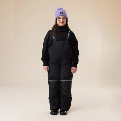 Salopette de neige Maxifit - Nanö noire, tissu imperméable avec renforts aux genoux, portée par enfant fille vue de face, style décontracté hiver.