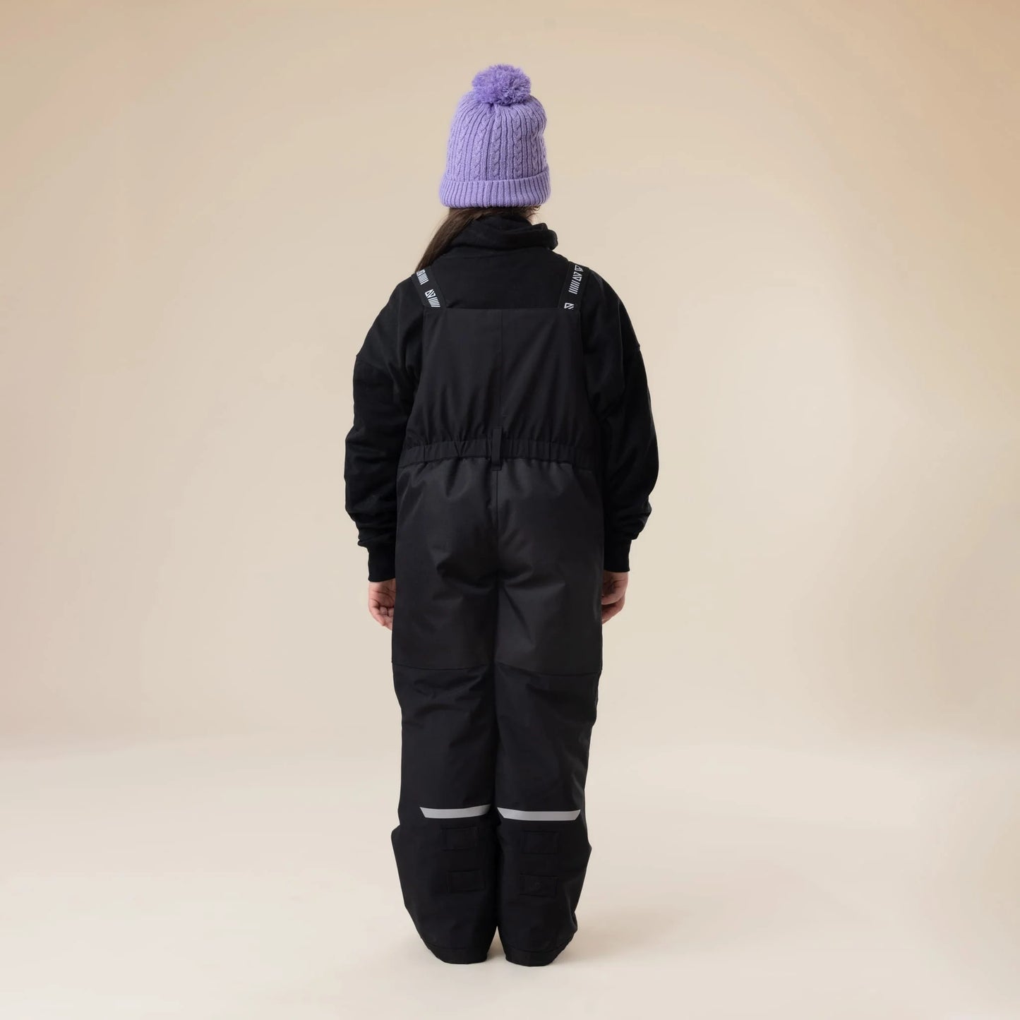 Salopette de neige Maxifit - Nanö noire en tissu imperméable, portée de dos par enfant, avec bonnet violet, style outdoor hiver.