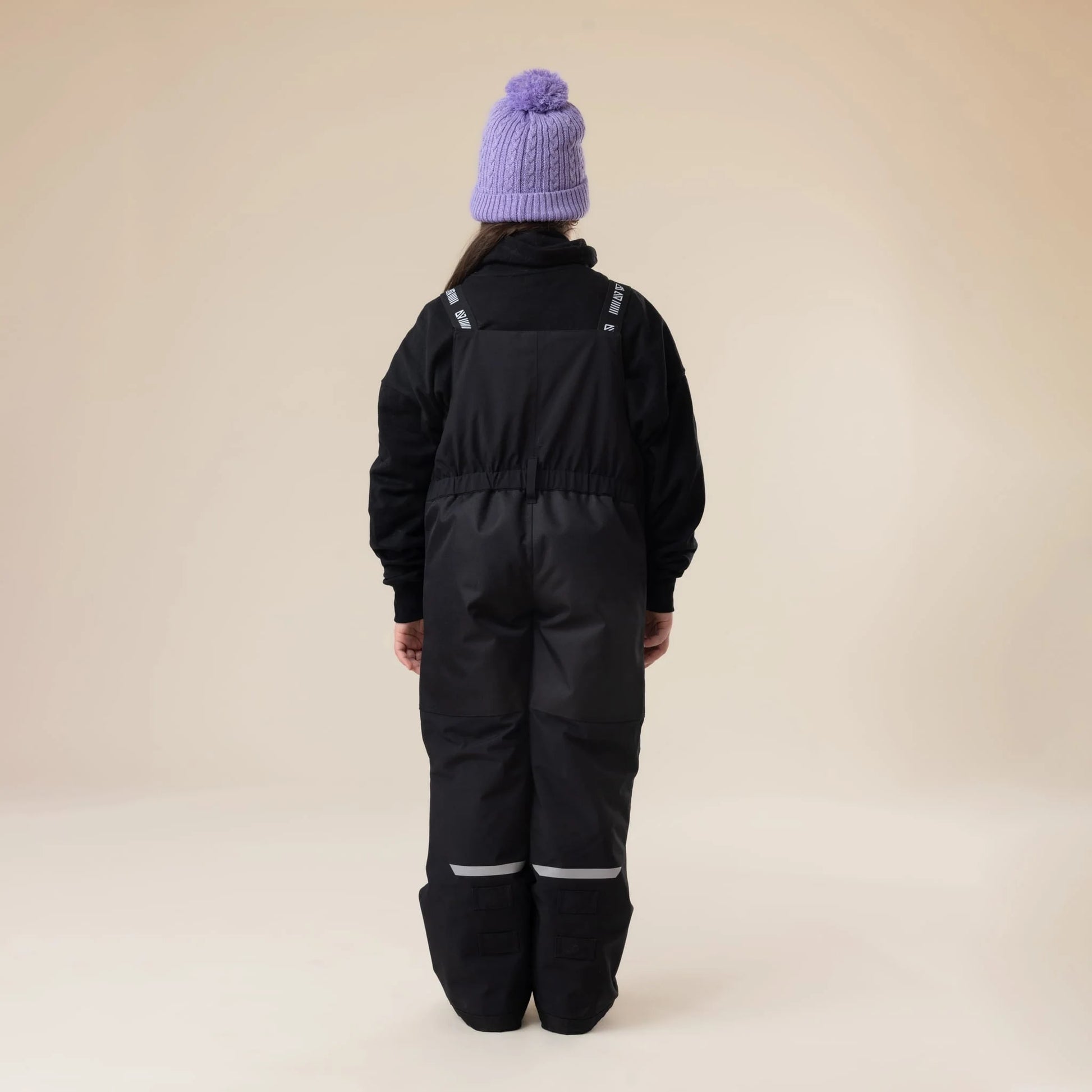Salopette de neige Maxifit - Nanö noire en tissu imperméable, portée de dos par enfant, avec bonnet violet, style outdoor hiver.