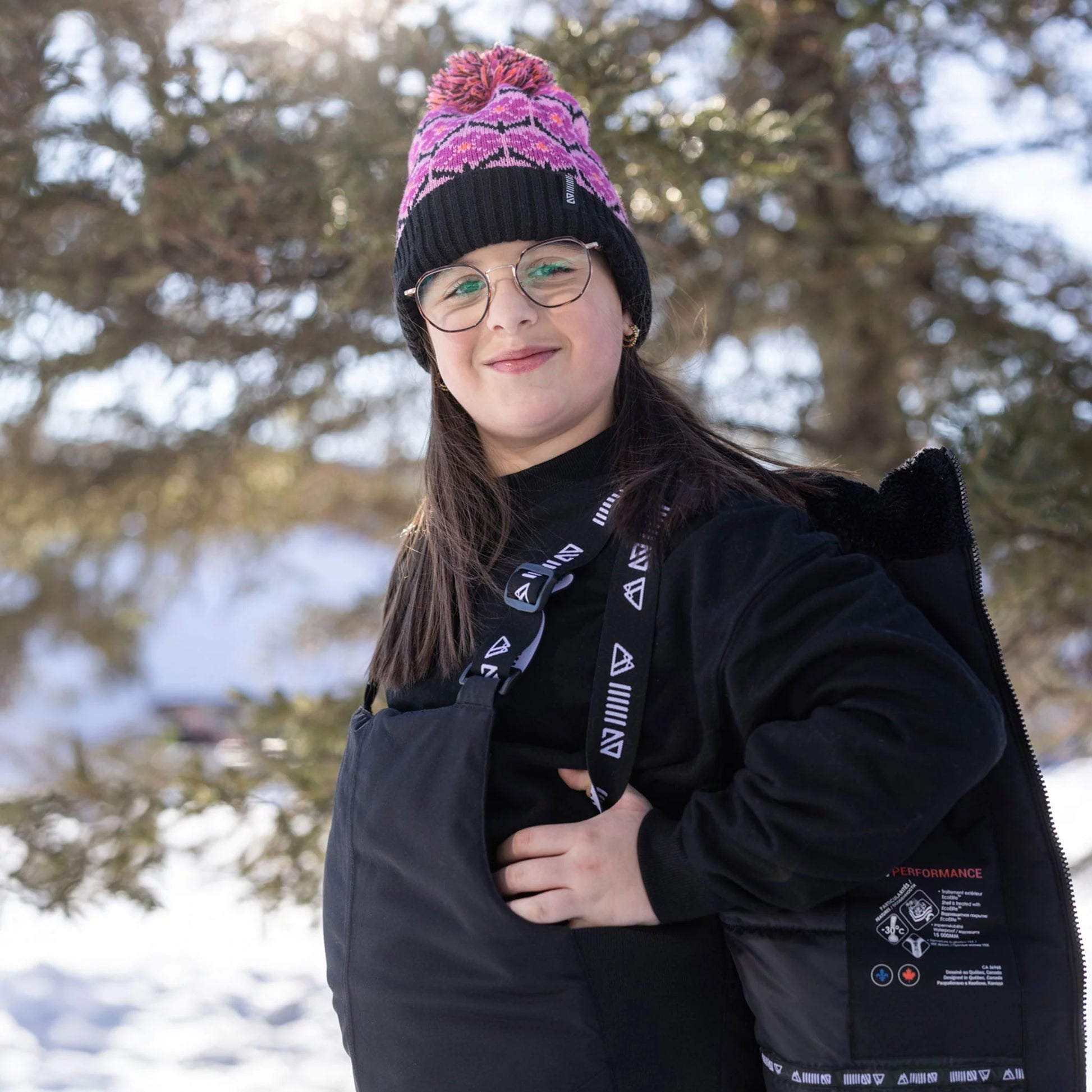 Salopette de neige Maxifit - Nanö noire en textile technique imperméable pour enfant, portée de face avec bonnet rose et noir en extérieur enneigé.