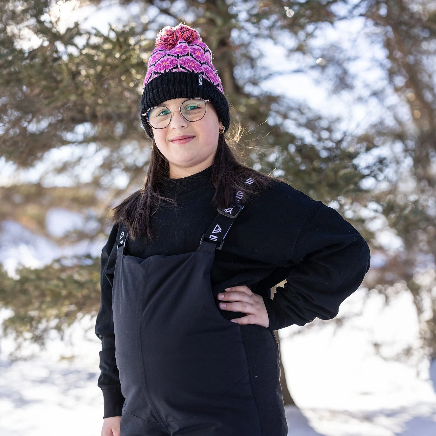 Salopette de neige Maxifit - Nanö noire portée par une enfant, vue de face, avec bonnet rose et noir, en extérieur enneigé.