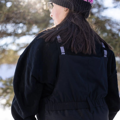 Salopette de neige Maxifit - Nanö noire en tissu imperméable, portée de dos par enfant avec bonnet, en extérieur neigeux.