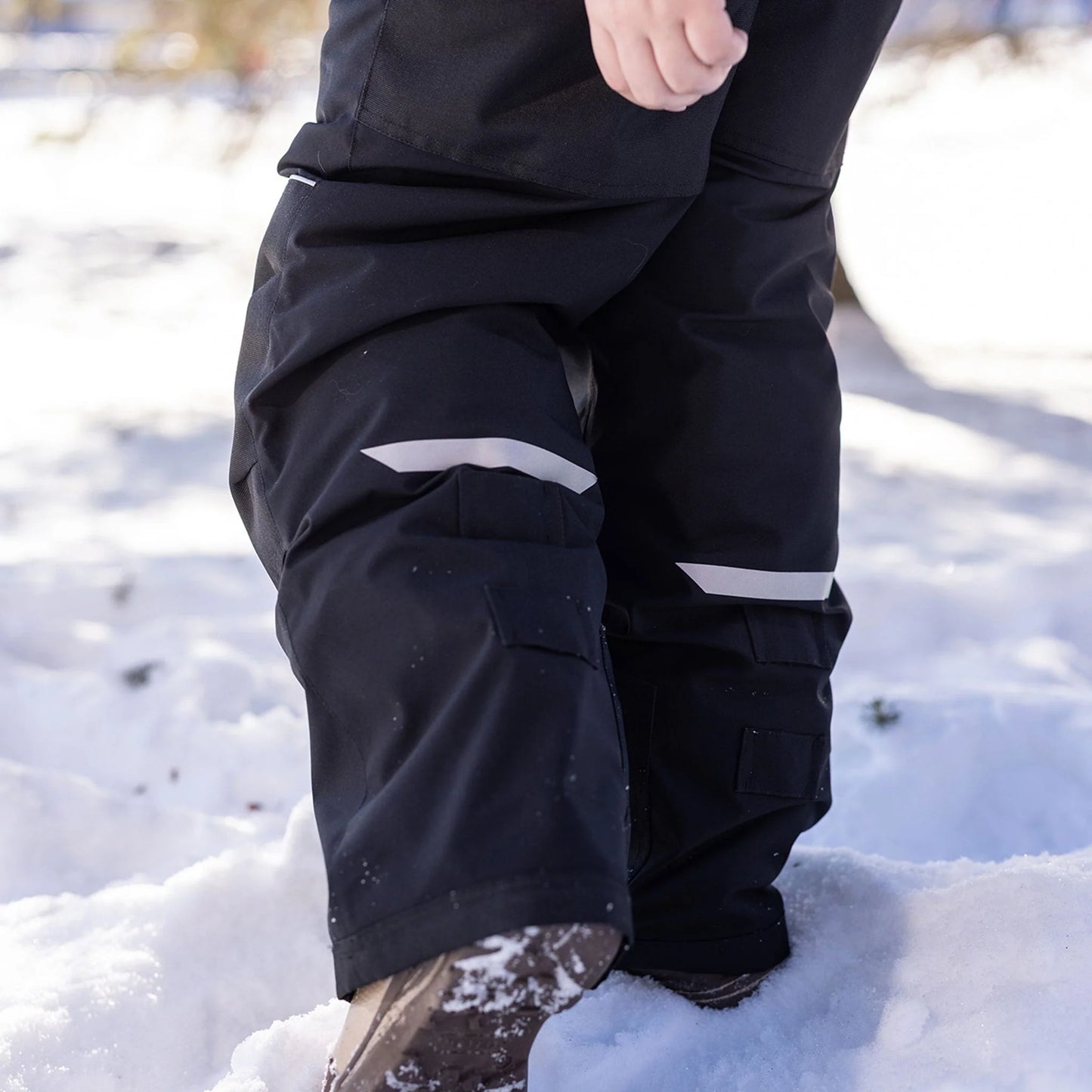 Salopette de neige Maxifit - Nanö noire avec bandes réfléchissantes grises, isolante pour enfant, vue en gros plan sur les jambes dans la neige.