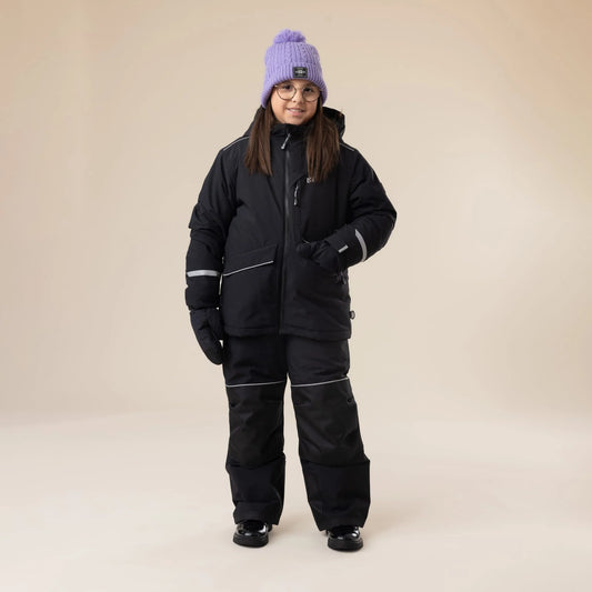Salopette de neige Maxifit - Nanö noire avec détails réfléchissants, portée par enfant fille vue de face avec bonnet violet et lunettes.