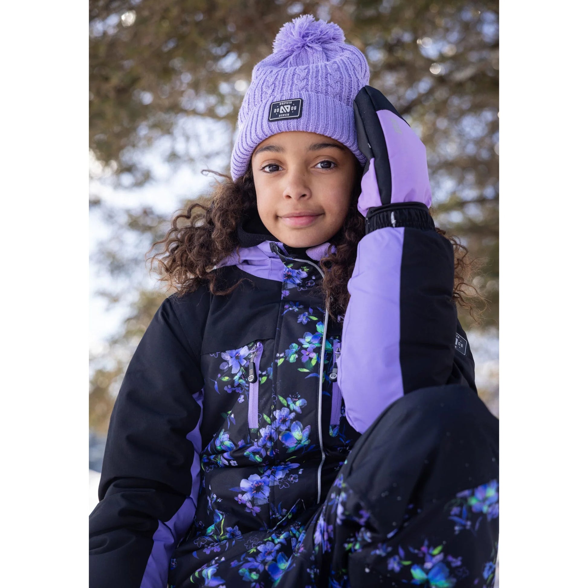 Habit de neige 1 pièce Emma - Nanö, combinaison noire et violet avec imprimé floral, portée par une enfant, vue de face en extérieur d’hiver.