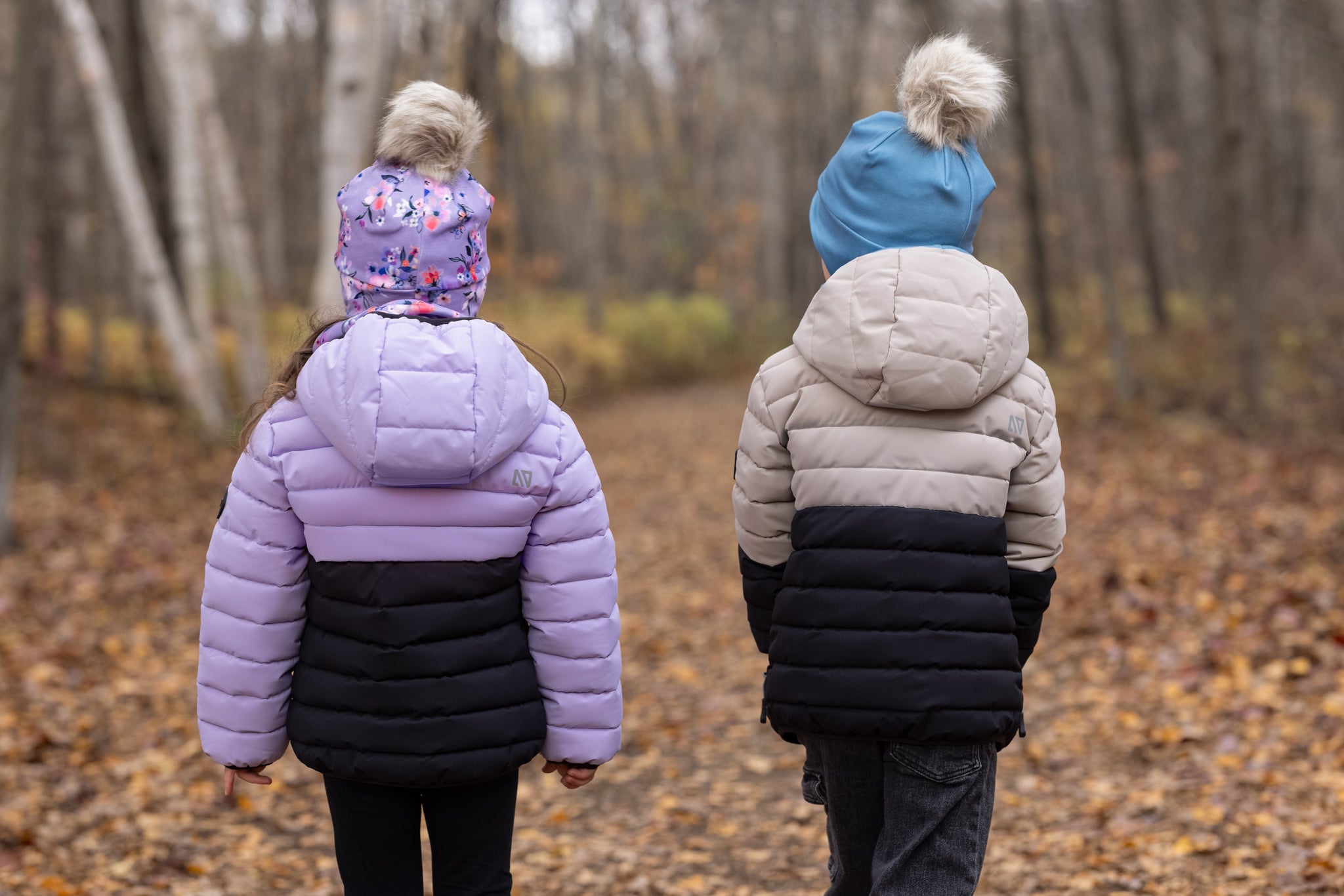Doudoune matelassée bicolore lilas et noir pour enfant fille à gauche, beige et noir pour enfant garçon à droite, vue de dos, avec bonnets à pompon en forêt automnale.